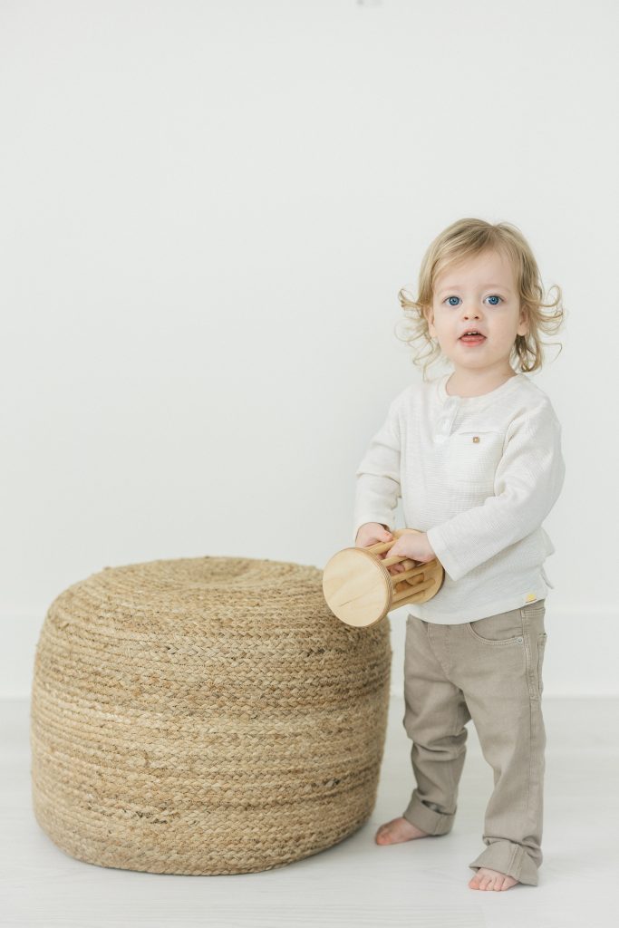 2 year old boy standing up next to a poof holding onto a wooden toy rattle in all white studio by NYC Family Photography