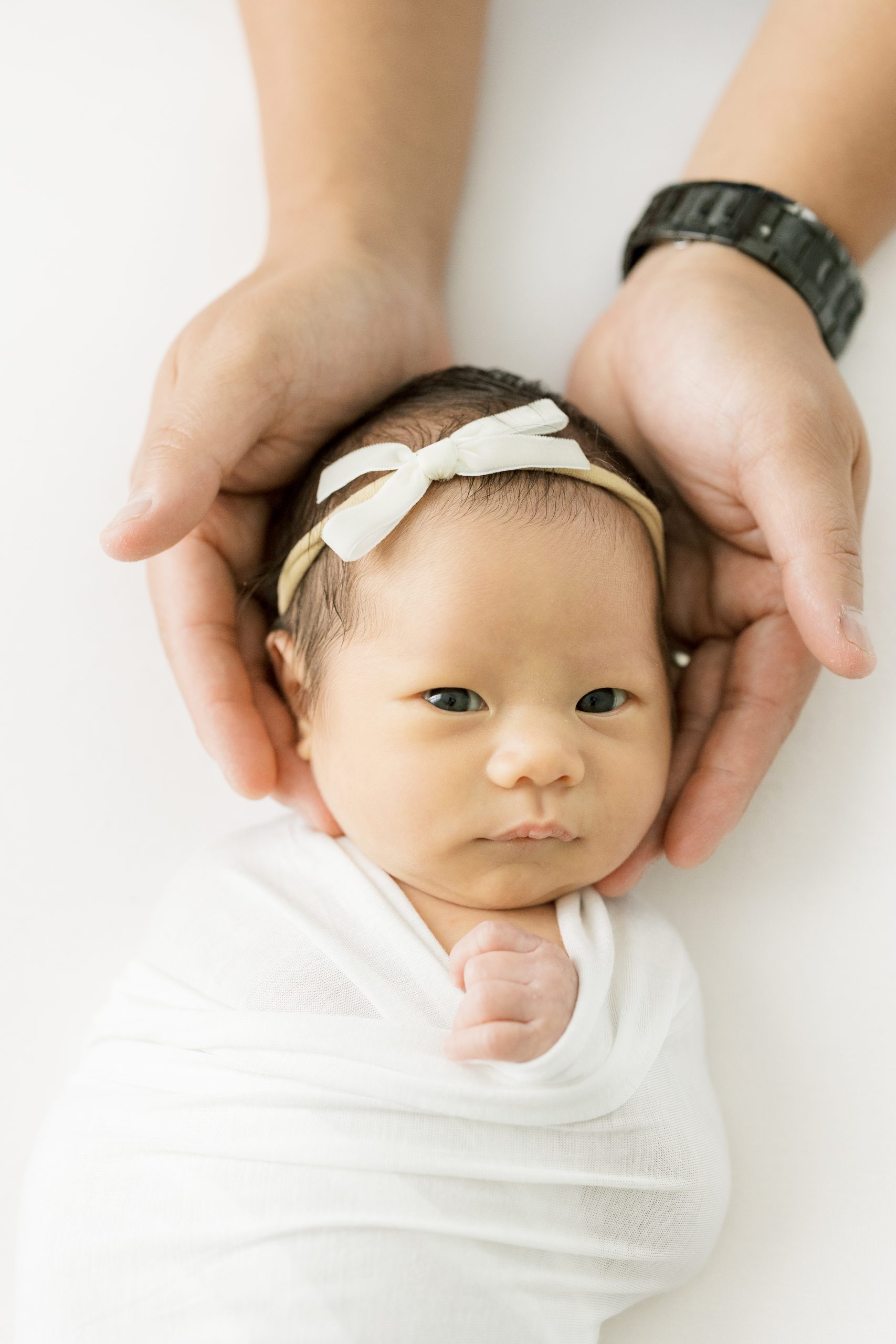 Newborn baby with white bow in her hair with her head being cradled by a set of hands in all white studio by NJ baby photographer