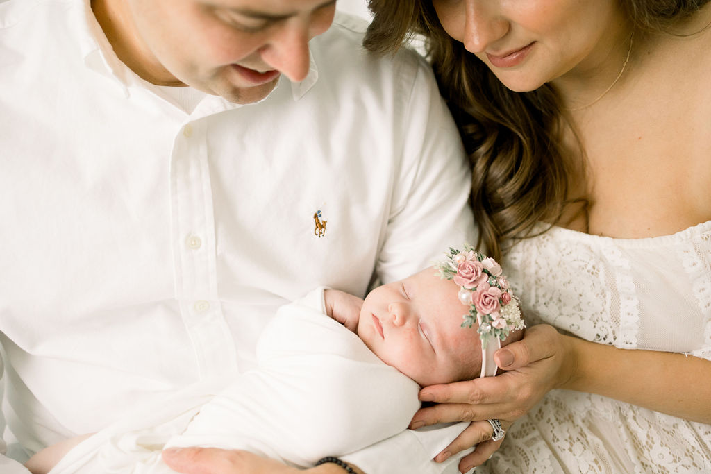 Mother and father look down on newborn baby girl in flower headband and white swaddle