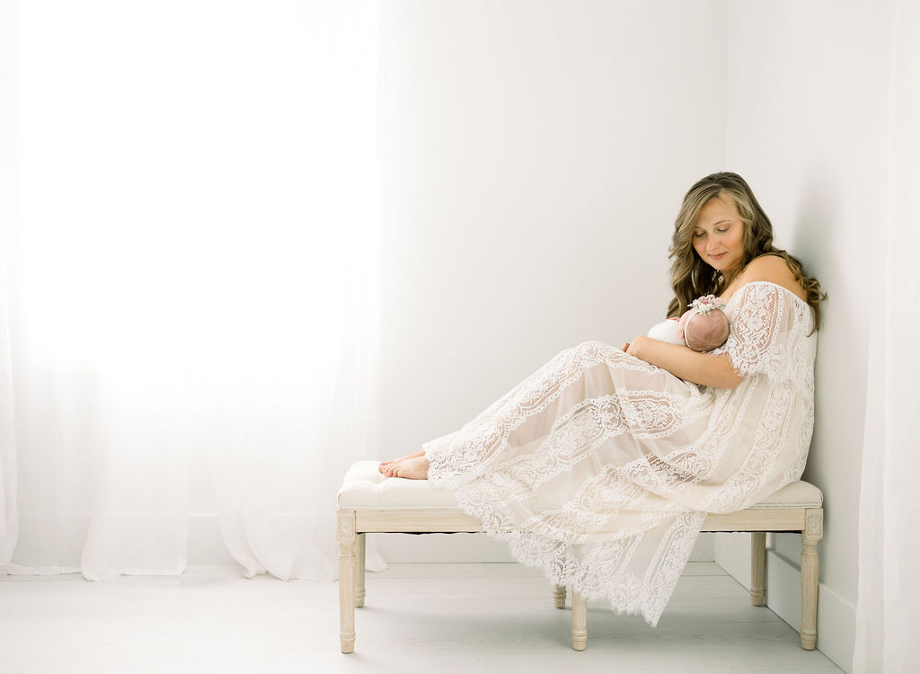 A mother in a white dress holds her newborn while sitting on a bench against a wall with her feet up