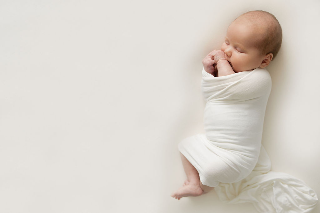 A newborn baby sleeps wrapped in a white blanket with feet and hands sticking out