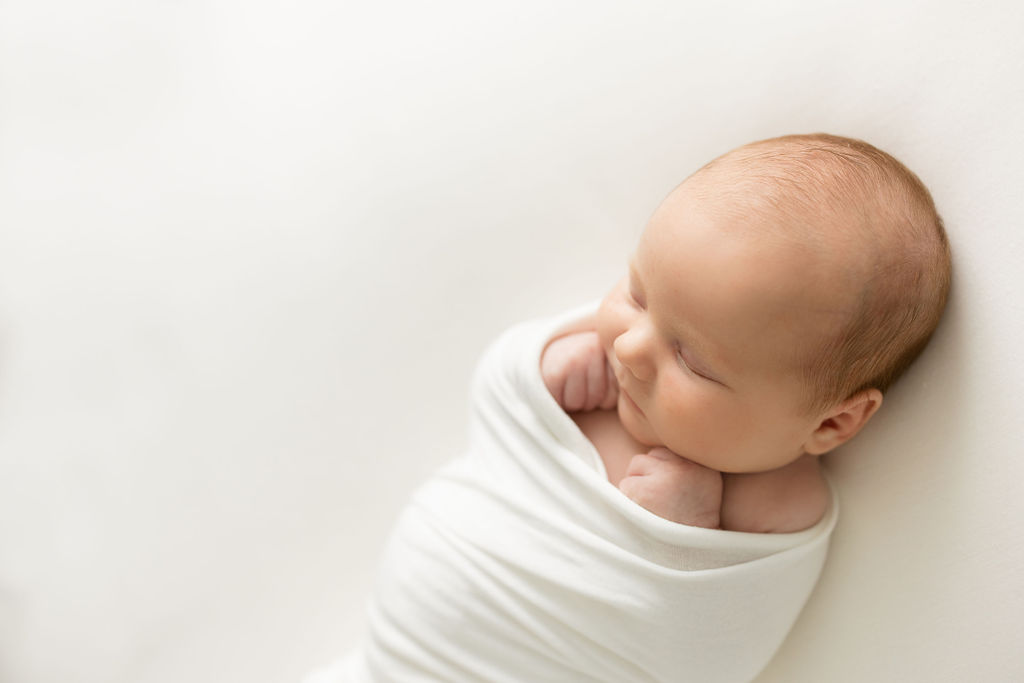 A newborn baby sleeps in a white swaddle on a white bed with hands in fists
