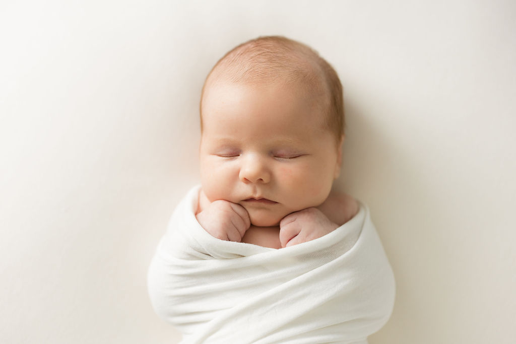 A newborn baby sleeps in a swaddle with hands under her chin on a white bed