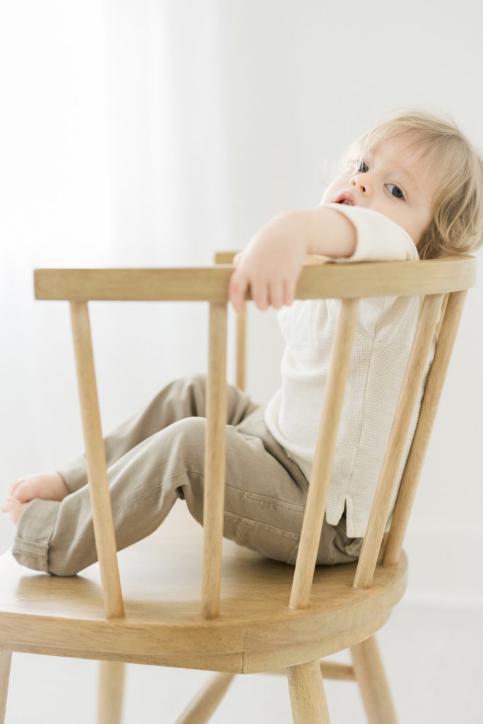 2 year old boy in warm white studio leaning his head back looking over his shoulder on a white oak chair by NYC Family Photography