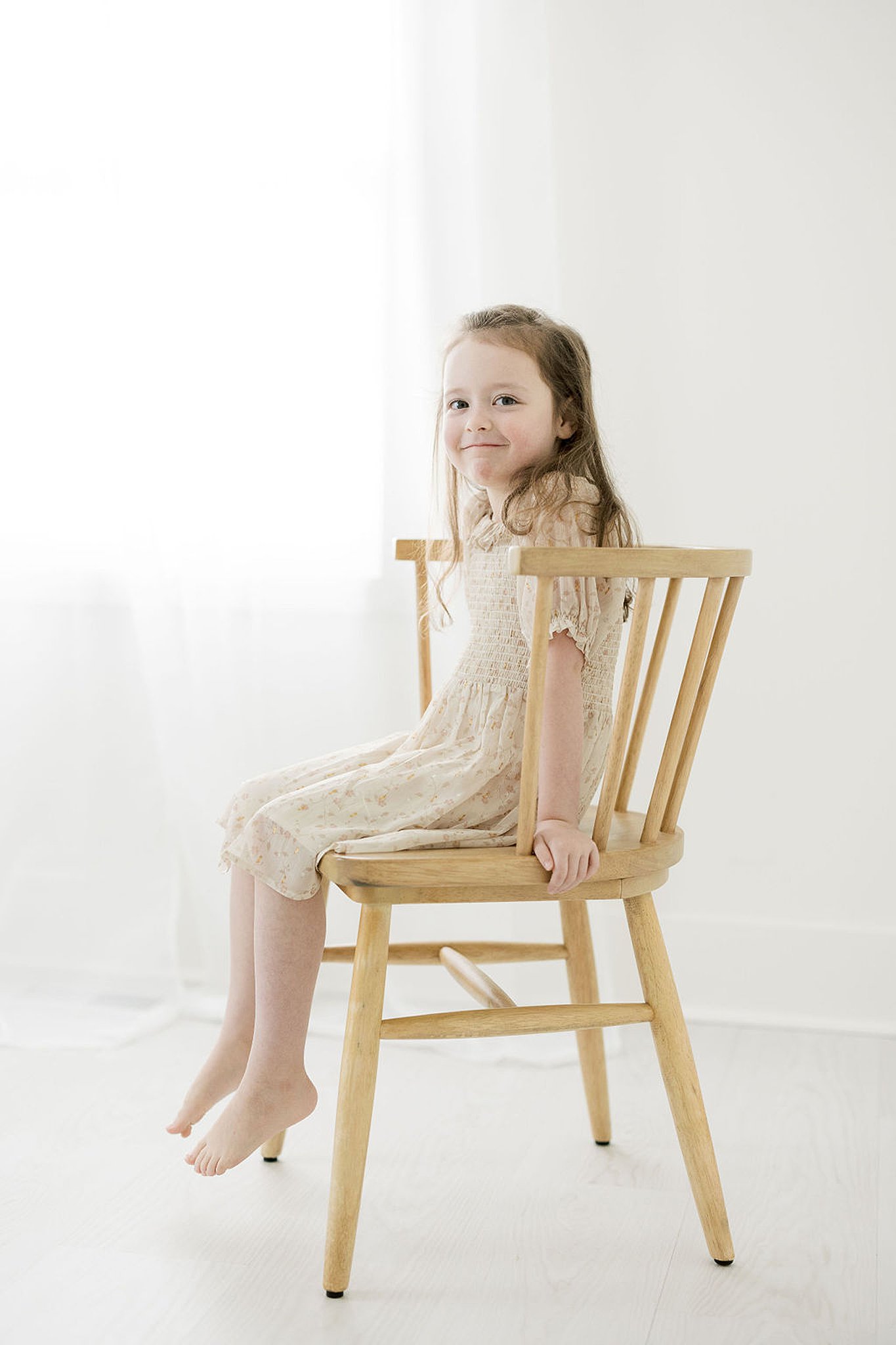 young girl sits on a tall wooden chair in an off-white dress in front of a window Different strokes for little folks