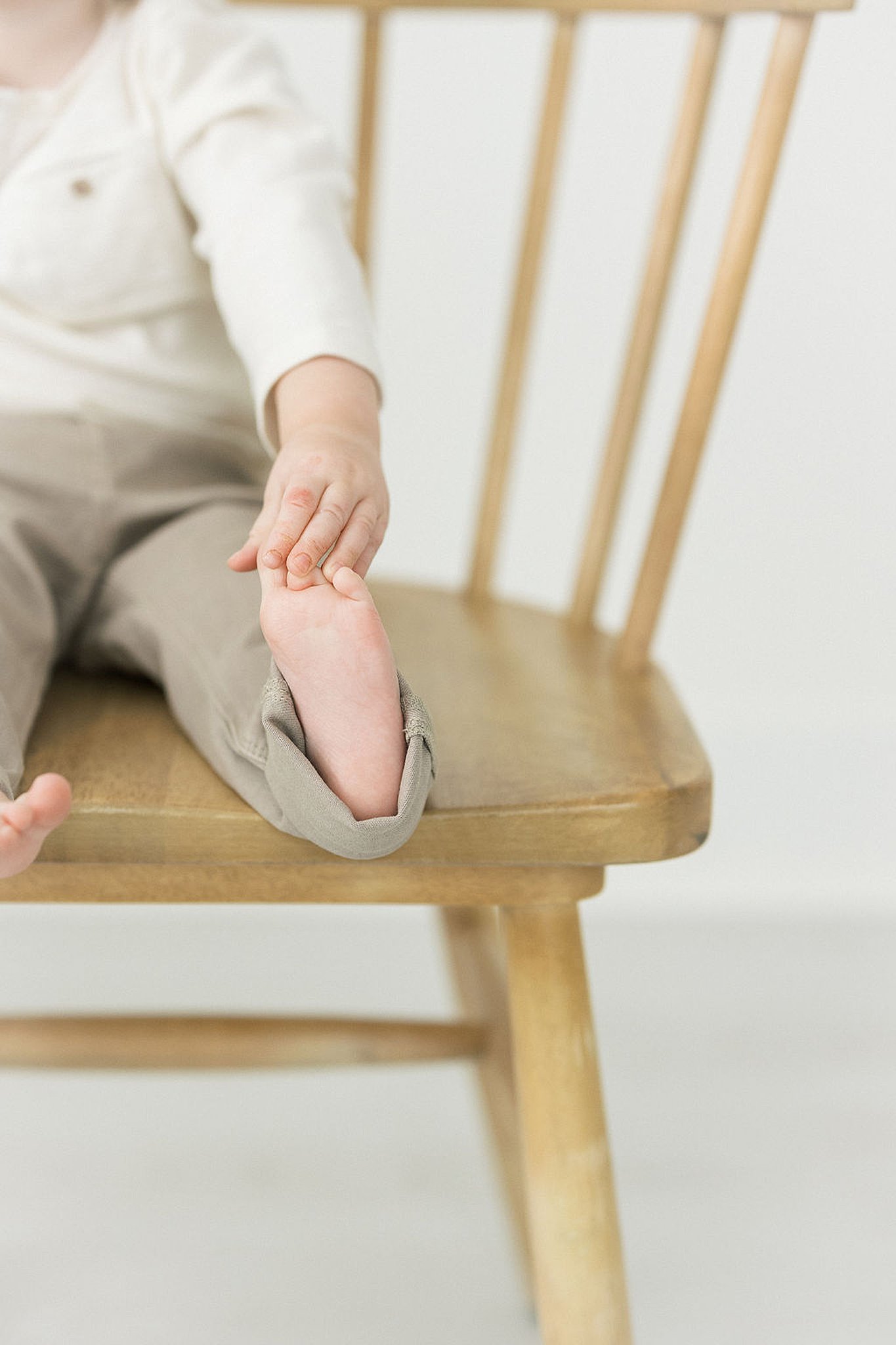 Toddler boy in khaki pants sits on a wooden chair holding his toes Hopscotch maplewood