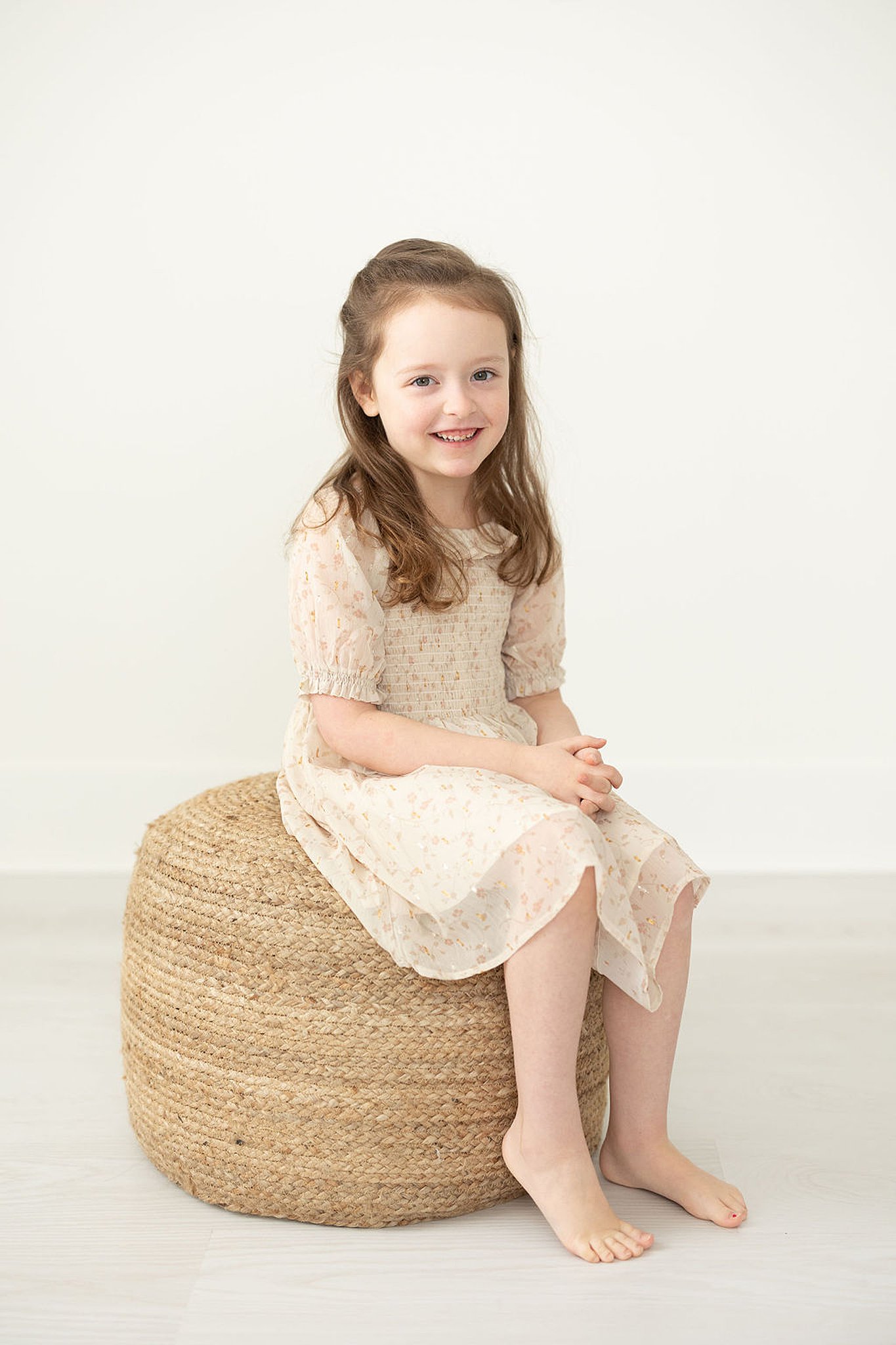 Girl in a patterned off-white dress sits on a woven stool in a studio Murray and finn