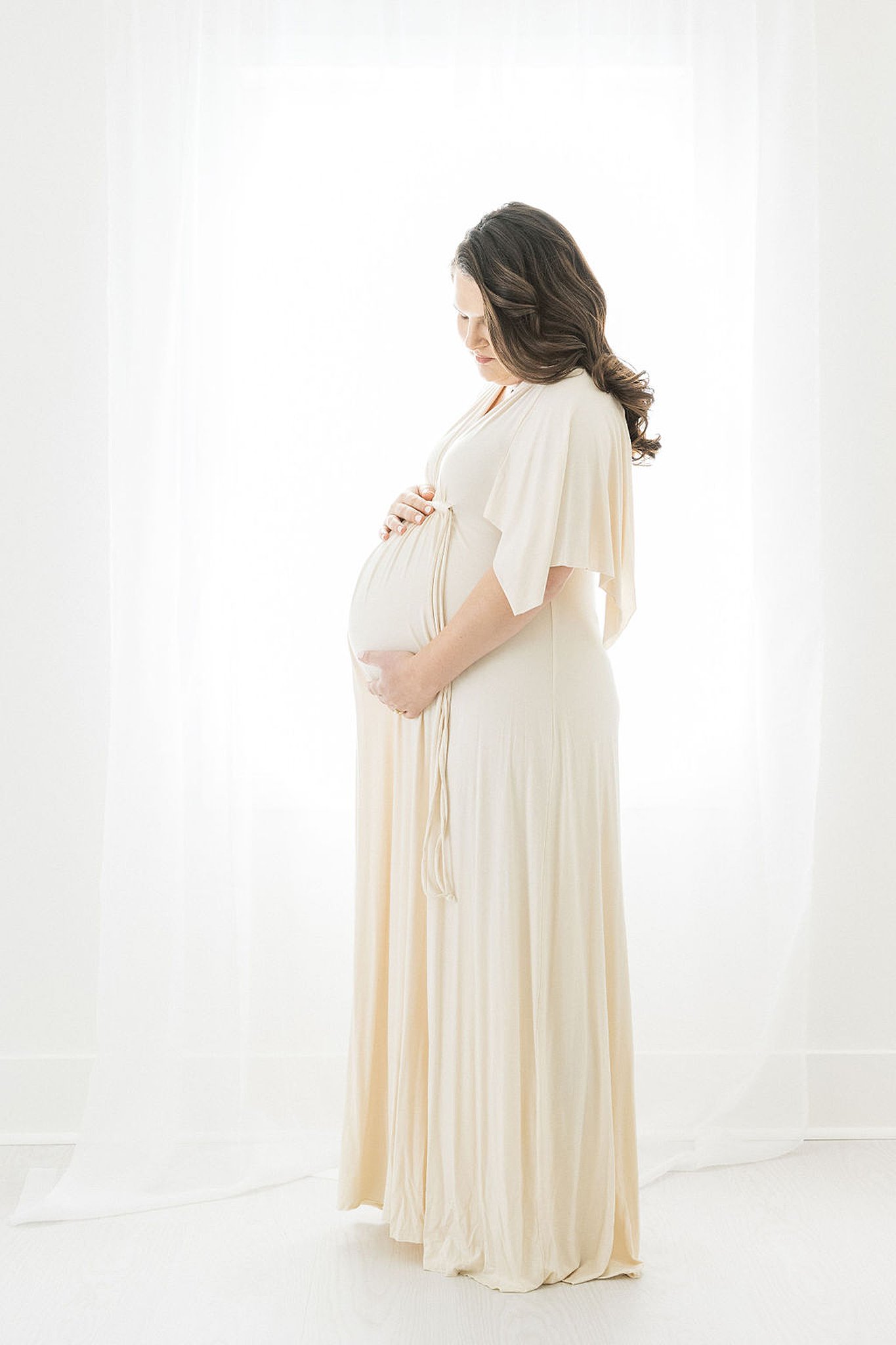 Mother-to-be stands in front of a window holding her bump with both hands and gazing down at it