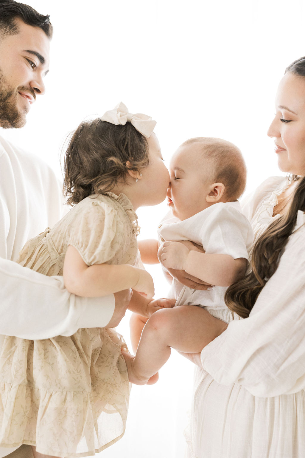 Toddler girl in a beige dress kisses her newborn sibbling's nose while both being held by parents