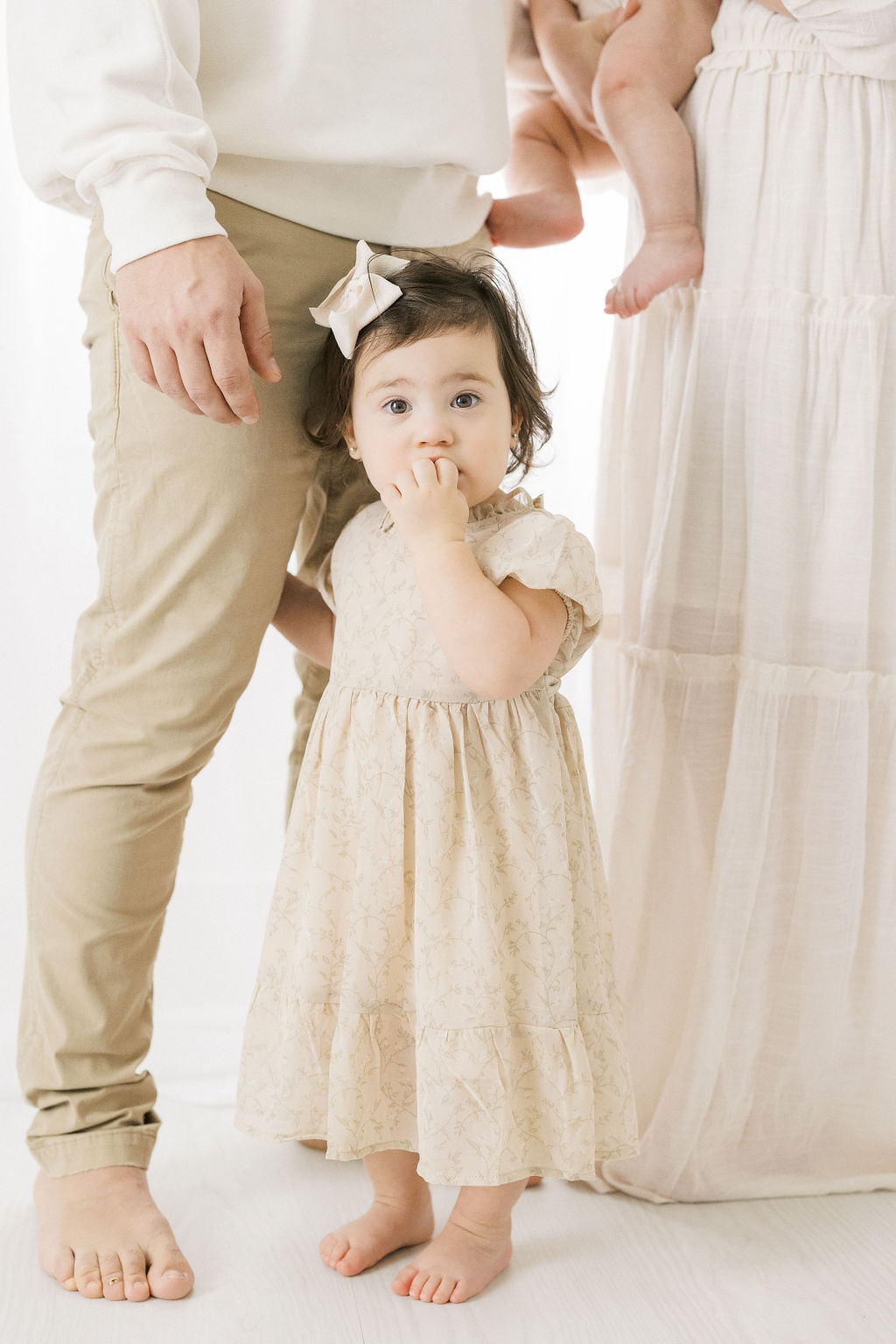 Toddler girl stands in a studio in a khaki dress holding her dad's leg bambi baby middletown