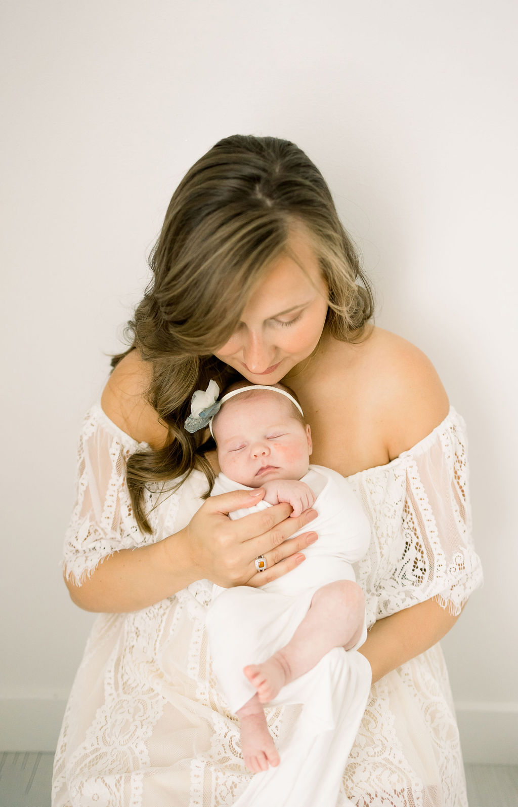 A mother in a white lace dress her newborn daughter while she grips her finger new jersey perinatal associates