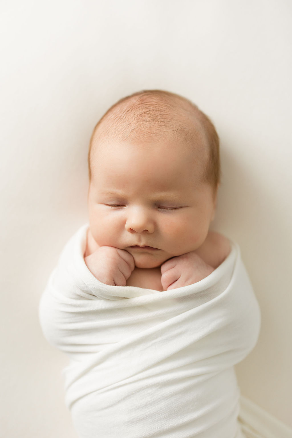 A newborn baby sleeps in a white swaddle on a white bed with hands in fists our birthing center