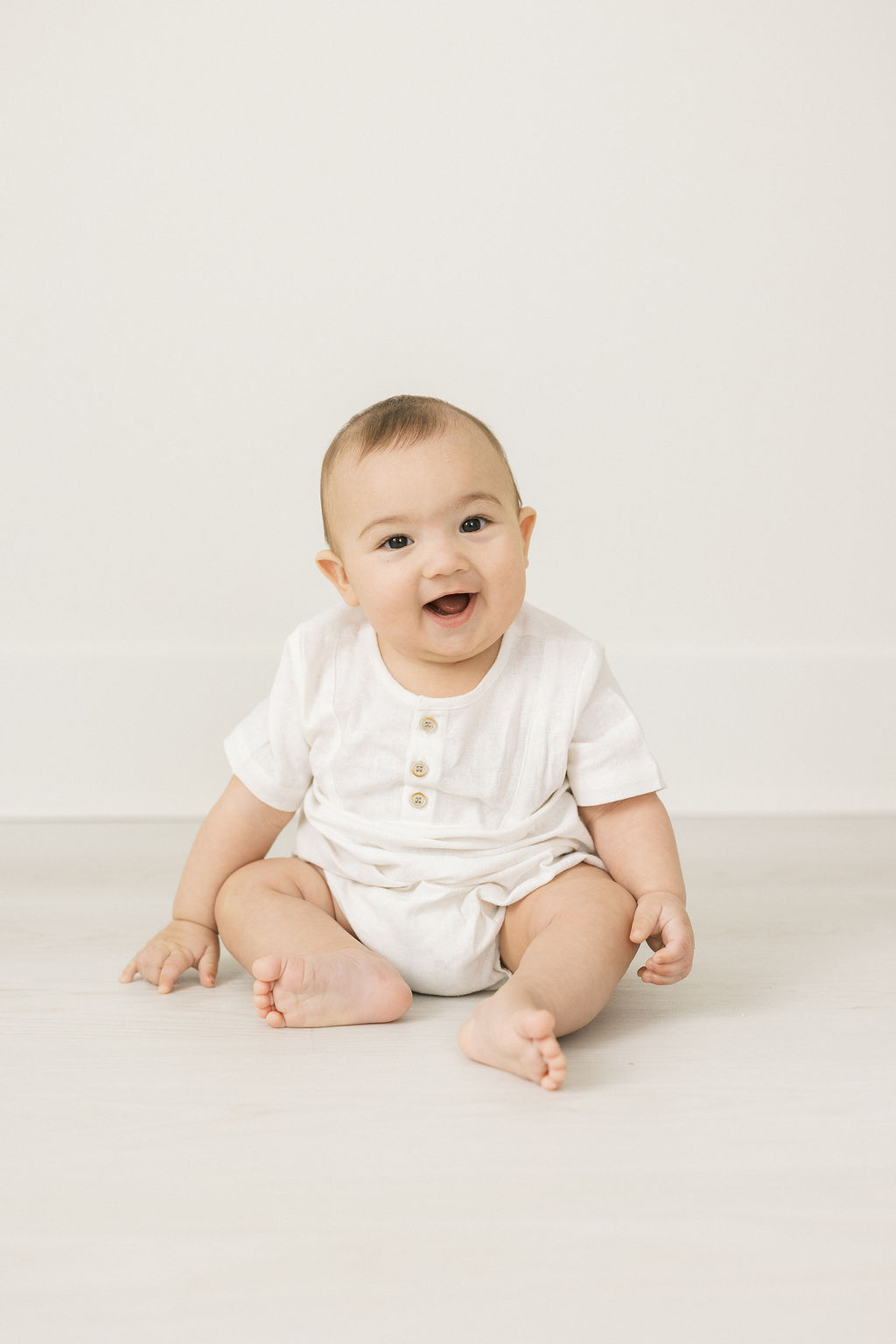 A toddler sits on a studio floor in a white onesie with buttons postpartum place chatham