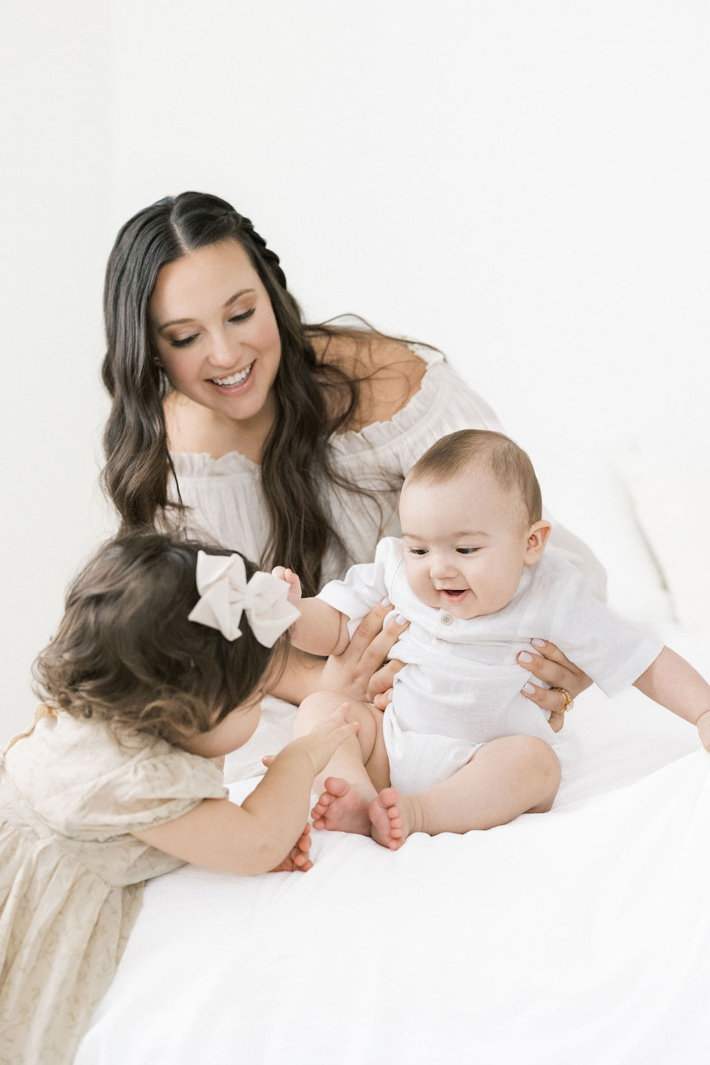 A toddler with dark hair plays with her newborn sibling and mom on a white bed postpartum place chatham