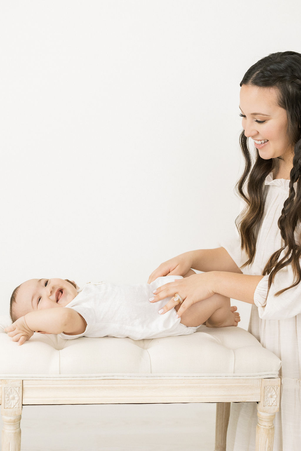 A mother tickles and plays with her toddler son on a beige bench wearing all white sage midwifery nj