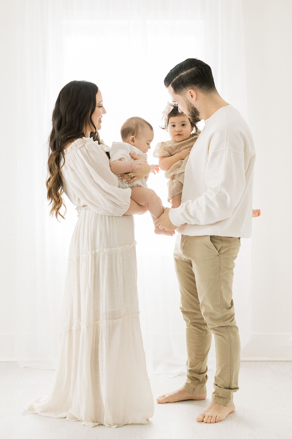 Parents in white stand in front of a window while holding their two toddler children in their arms