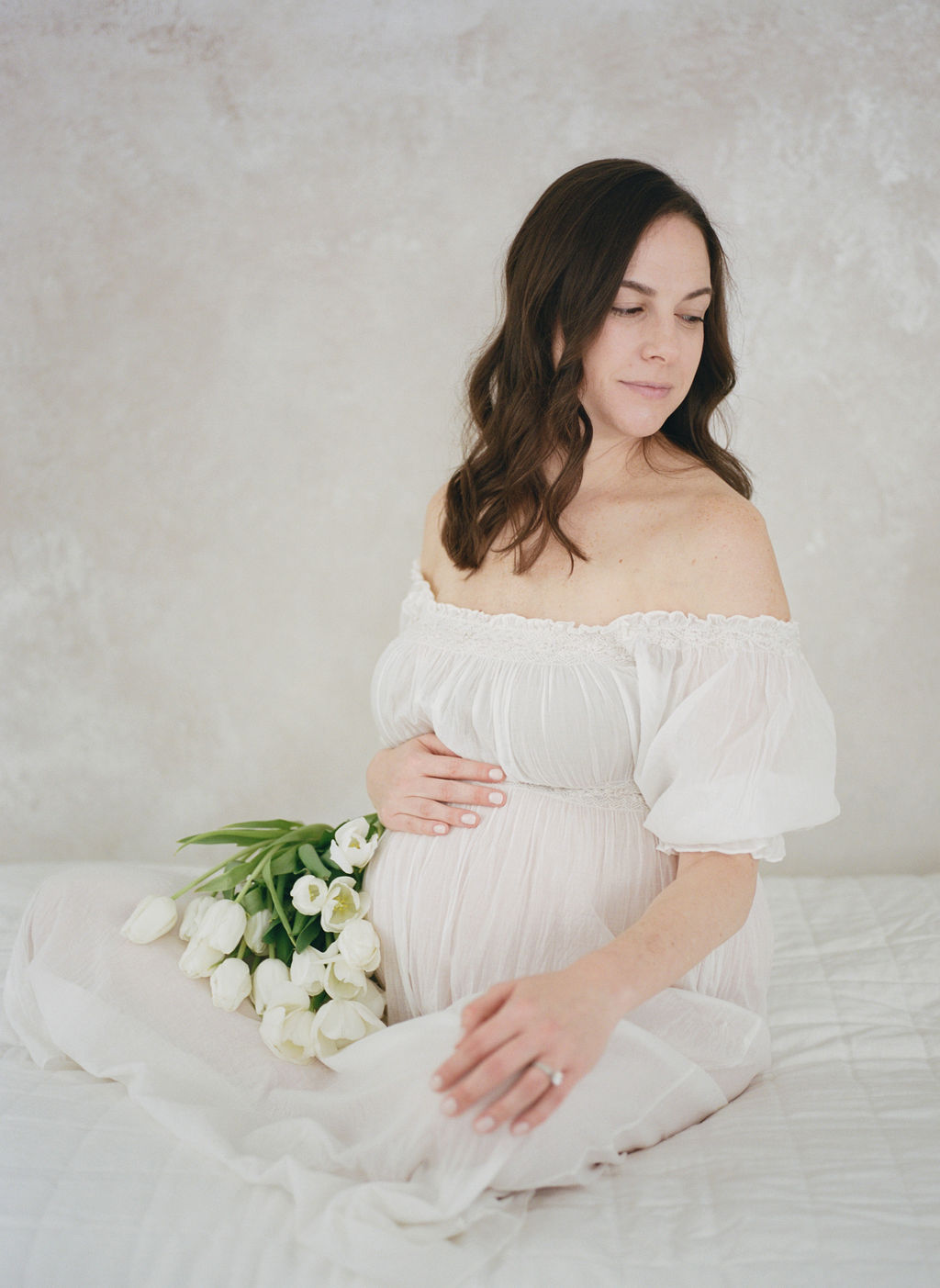 A mother to be in a white maternity dress sits with legs crossed on a white bed with white tulips in her lap childbirth center clifton