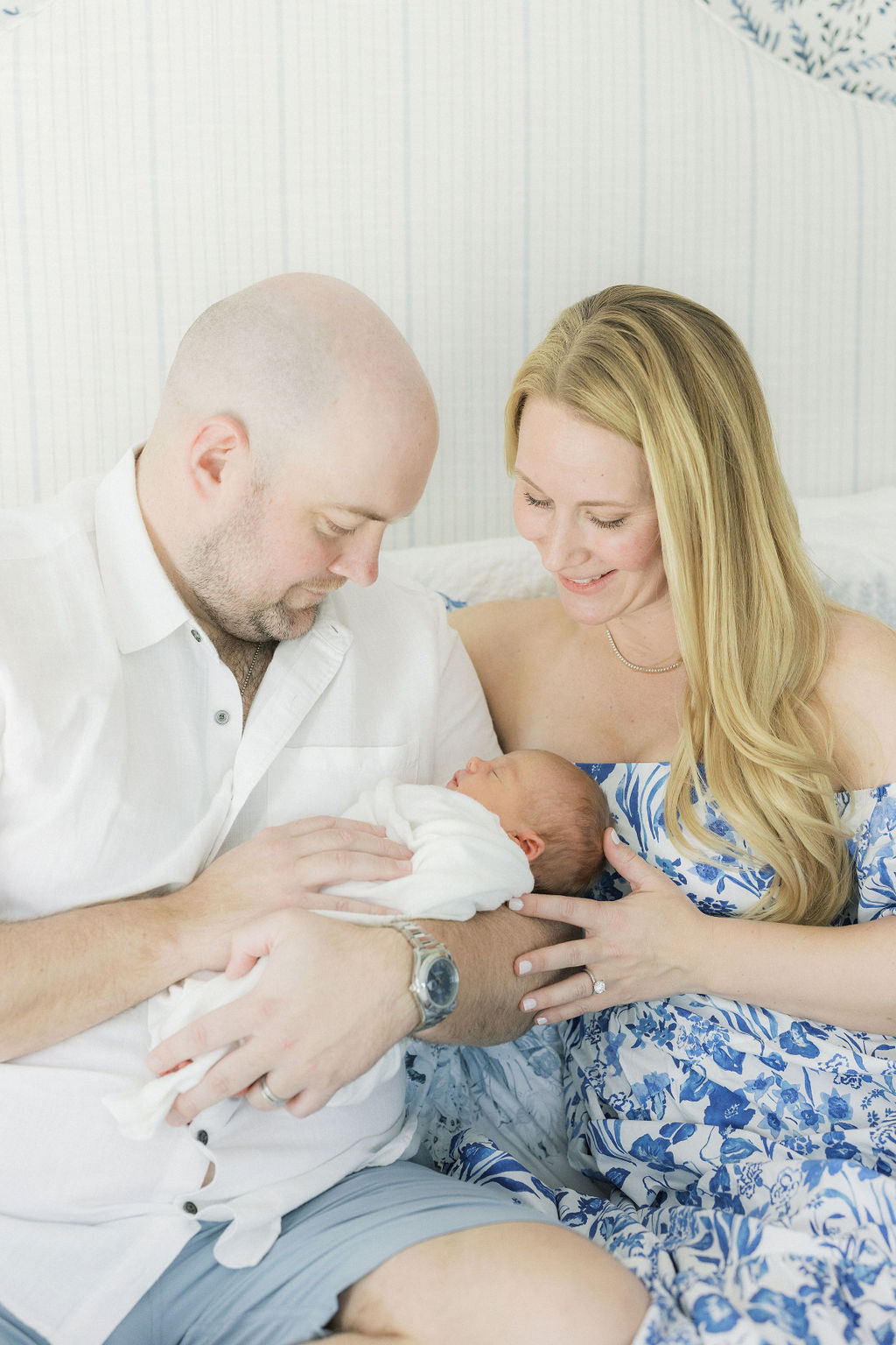 A father holds his newborn baby while sitting on a couch with mom in a blue dress leaning in next to them