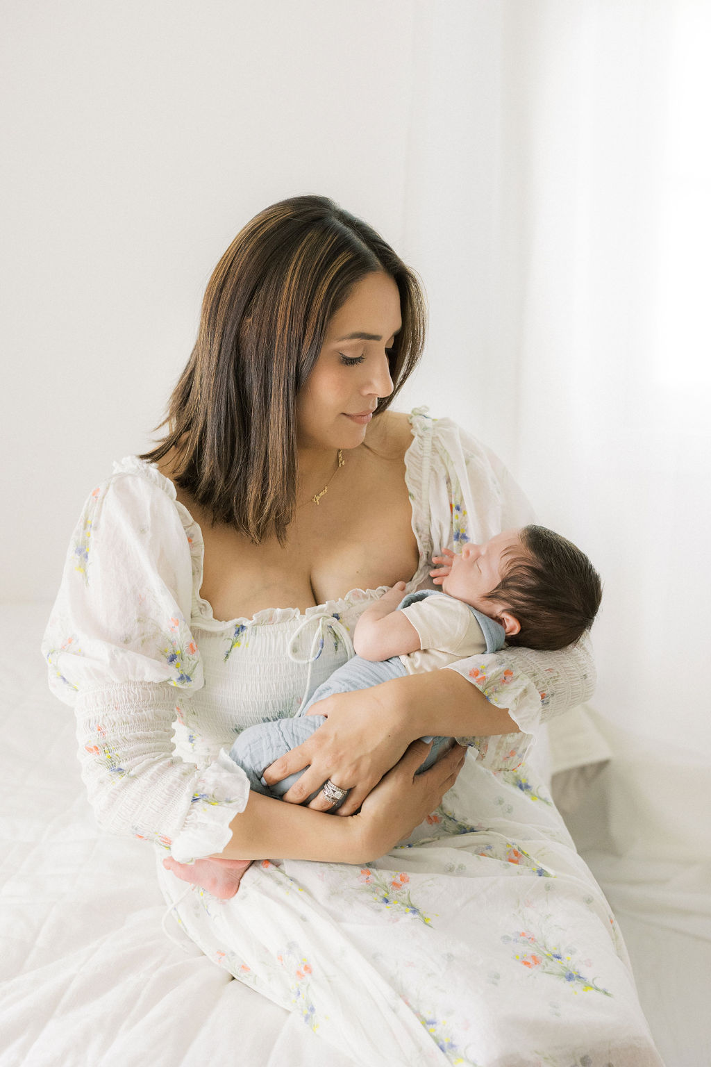 A mother sits on the edge of a bed holding her sleeping newborn baby boy in her arms