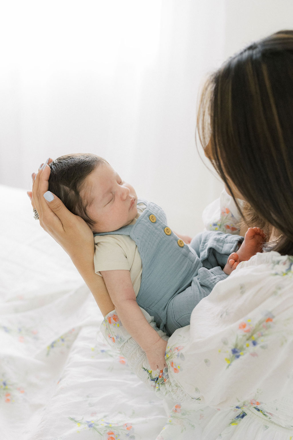 A mother holds her sleeping newborn baby boy in overalls facing her in front of a window laid back lactation