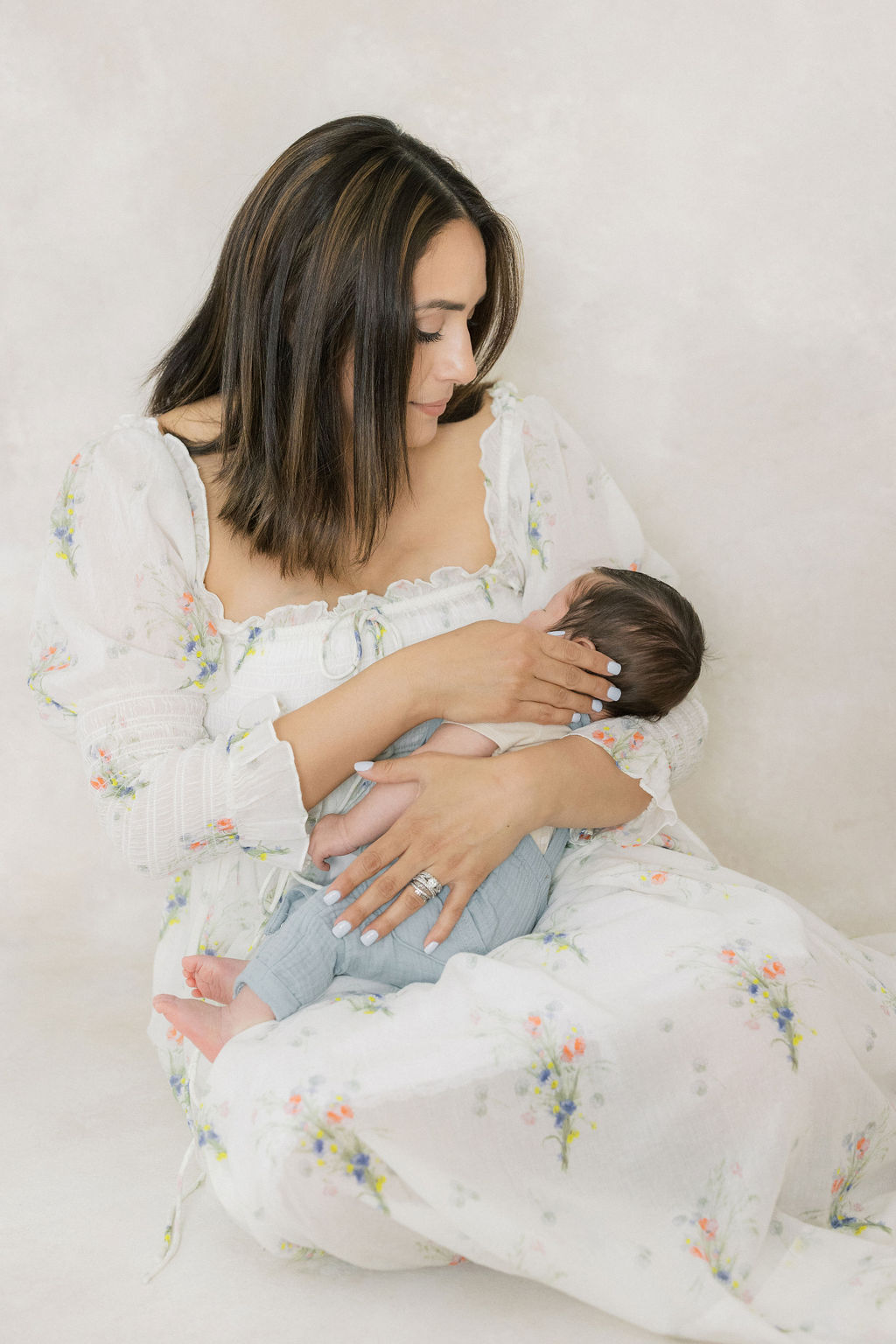 A new mother sits on the floor of a studio in a white dress with floral patterns holding her newborn baby boy in her lap laid back lactation