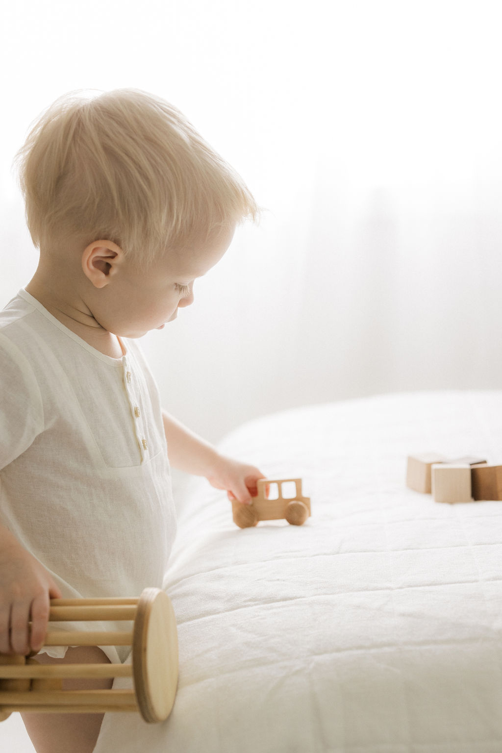A toddler boy in a white onesie plays with wooden toys on a white bed little ones livingston