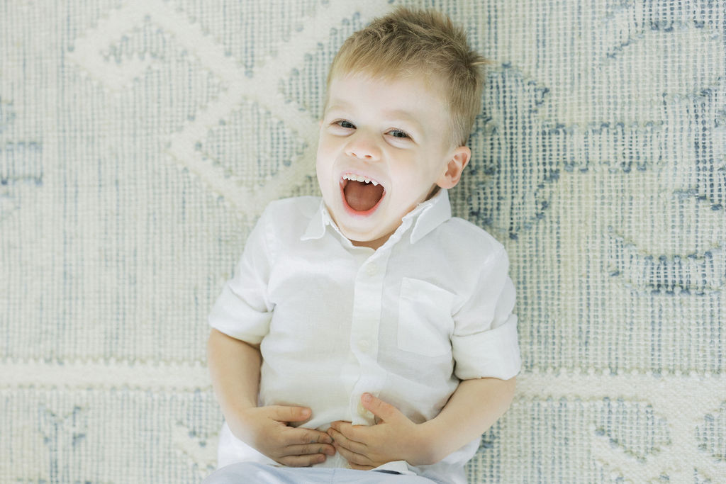 A young boy in a white button-down shirt lays on a rug with a huge smile