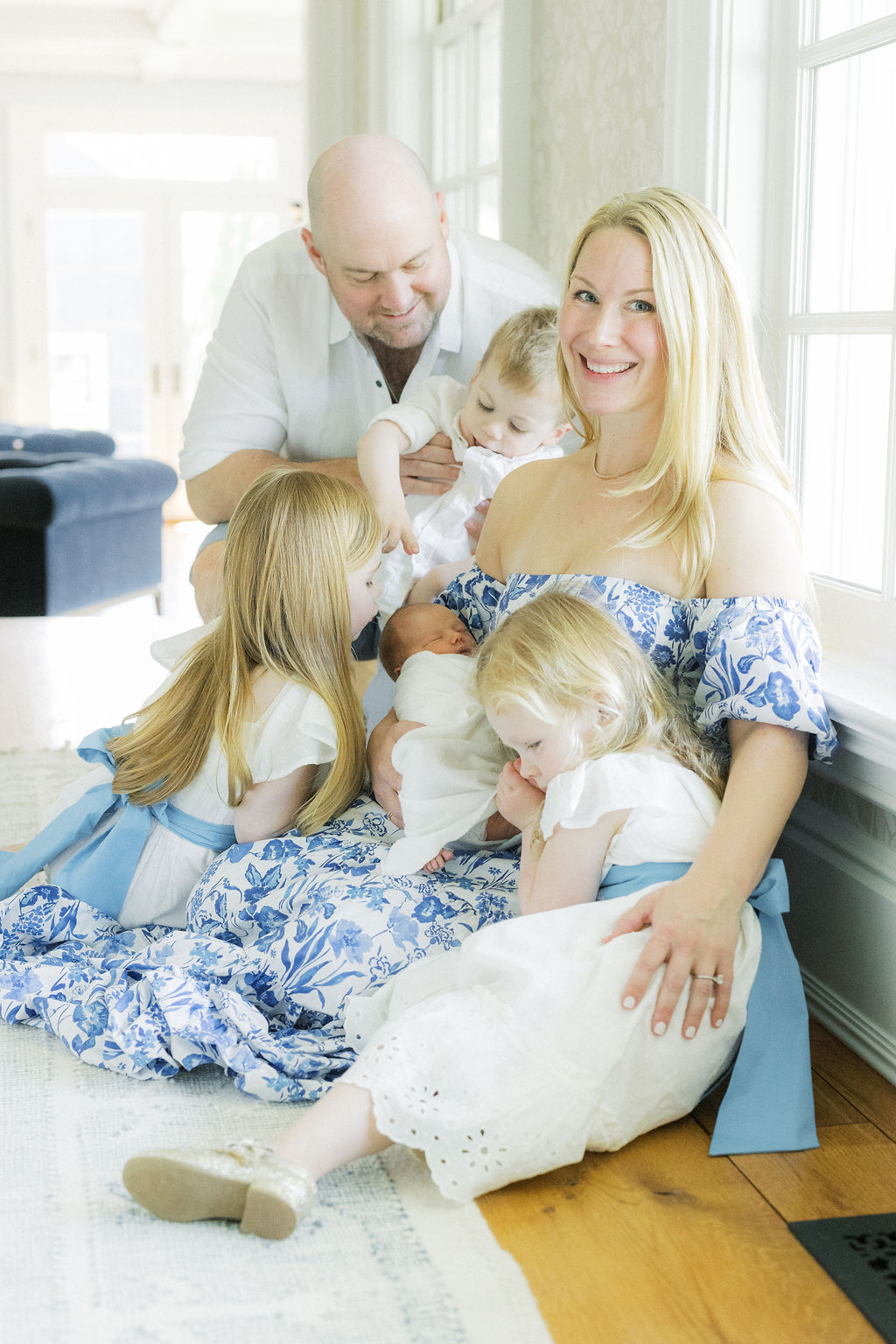 A mother sits by a window in a blue dress holding her newborn baby while her three other children and husband play and cuddle around her morristown daycare