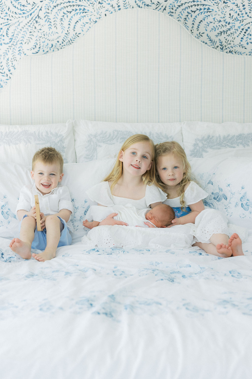 Three young siblings sit in a large bed while the oldest in the middle holds their newborn baby sibling morristown daycare