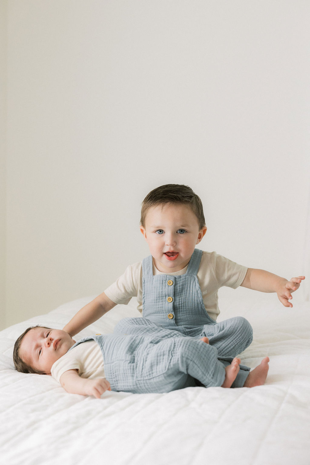 A toddler boy in blue overalls sits on a bed with his newborn baby brother laying in front of him