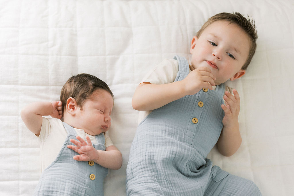 A newborn baby sleeps on a white bed while older brother lays next to him in matching blue overalls morristown pediatric dentist