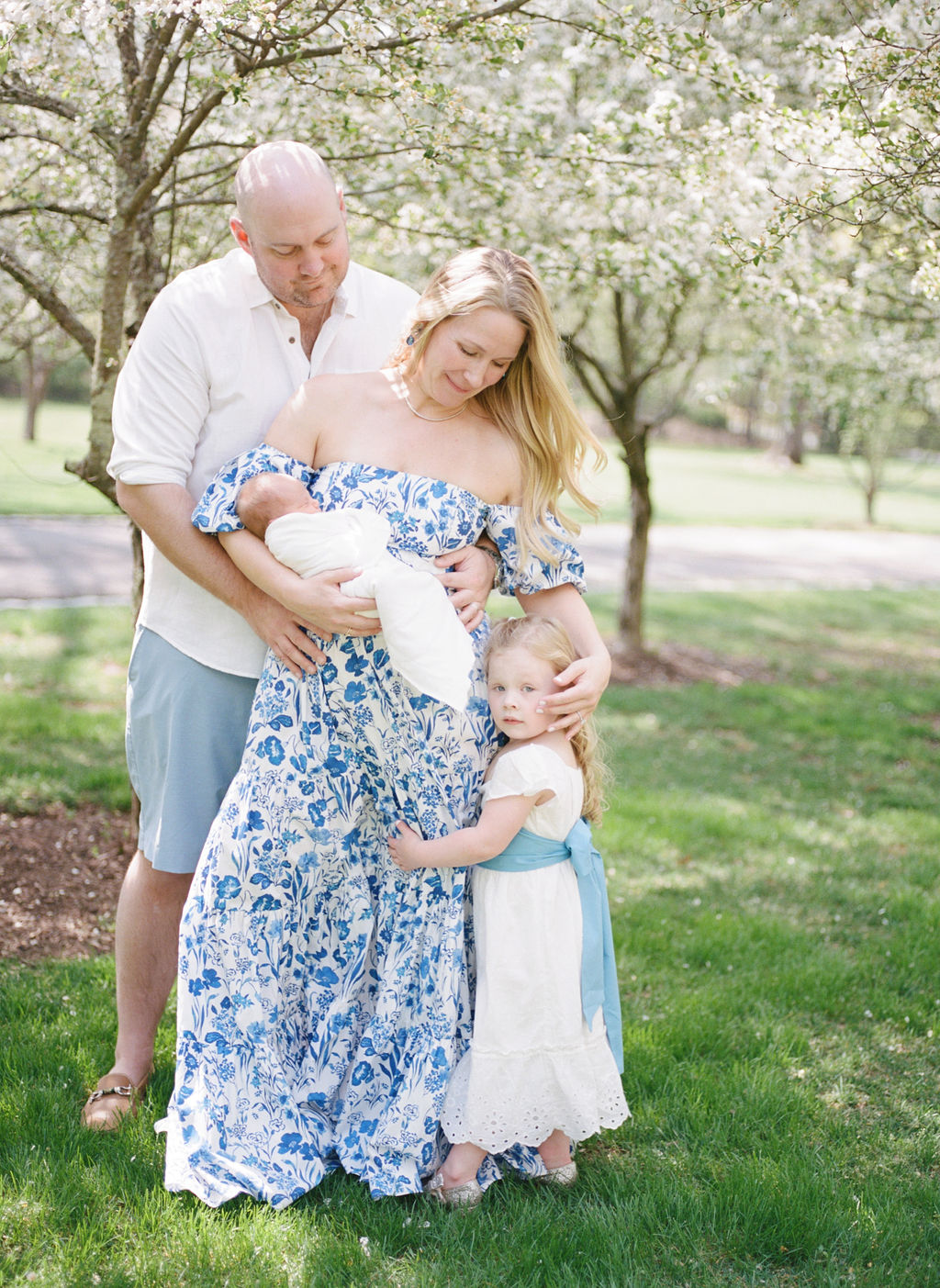 A mother stands in a park under blooming trees while holding her newborn baby with dad leaning over her shoulder and older daughter hugging her leg obgyn parsippany nj