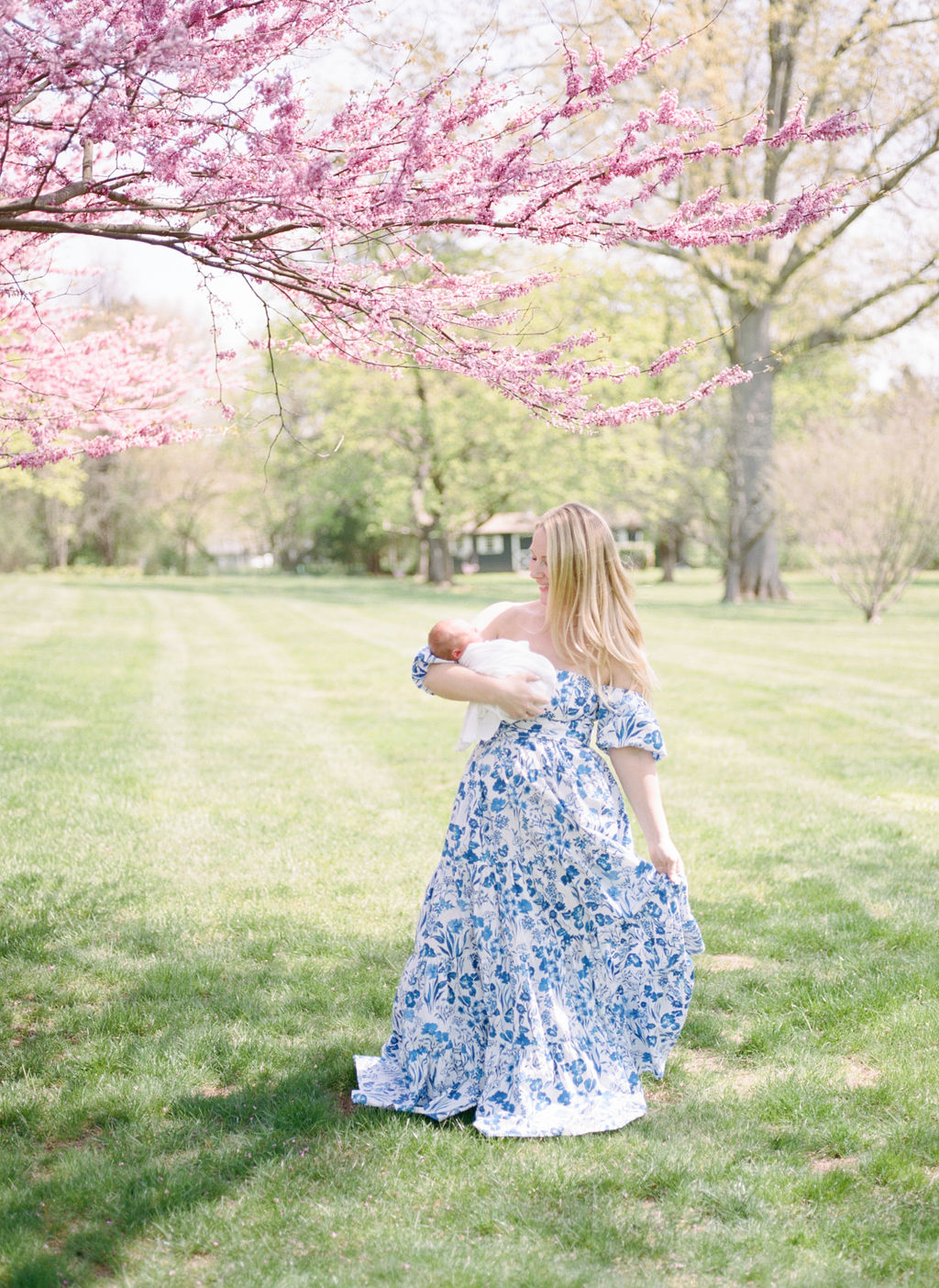 A mother in a blue dress dances in a grassy field under a pink dogwood tree while holding her newborn baby obgyn parsippany nj