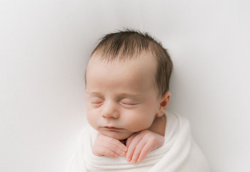 A newborn baby sleeps in a white swaddle with hands sticking out under its chin