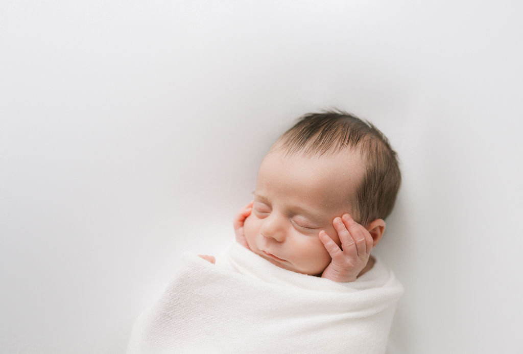 A newborn baby sleeps in a white swaddle with hands on cheeks overlook labor and delivery