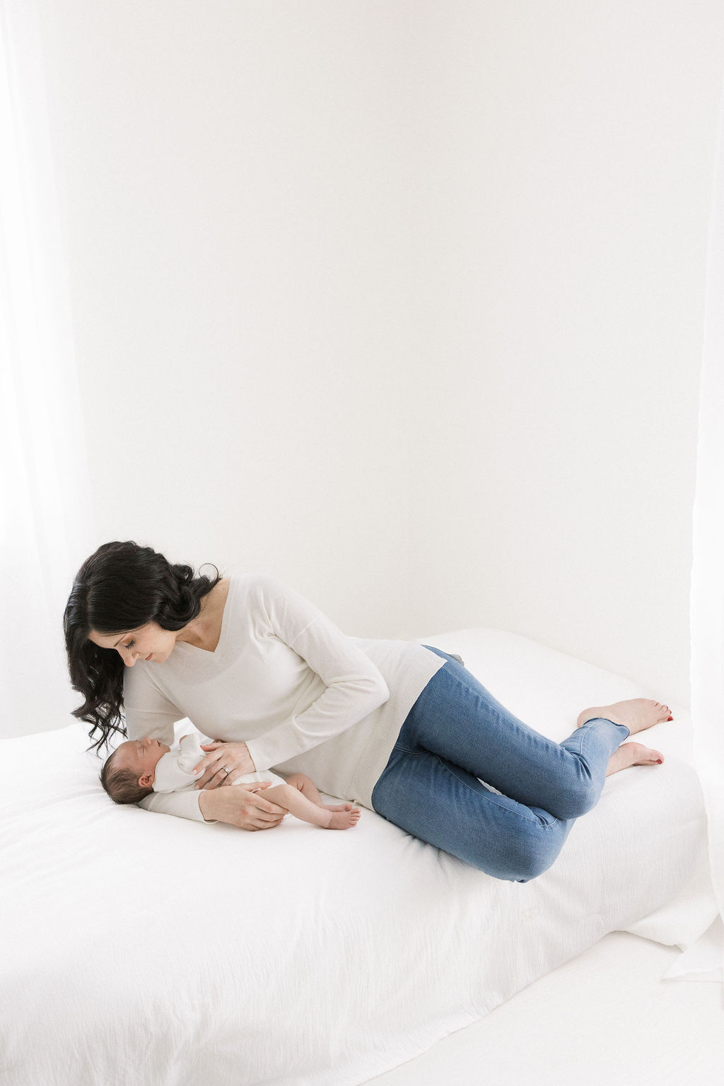 A mother in jeans lays across a bed with her newborn baby sleeping on her arm