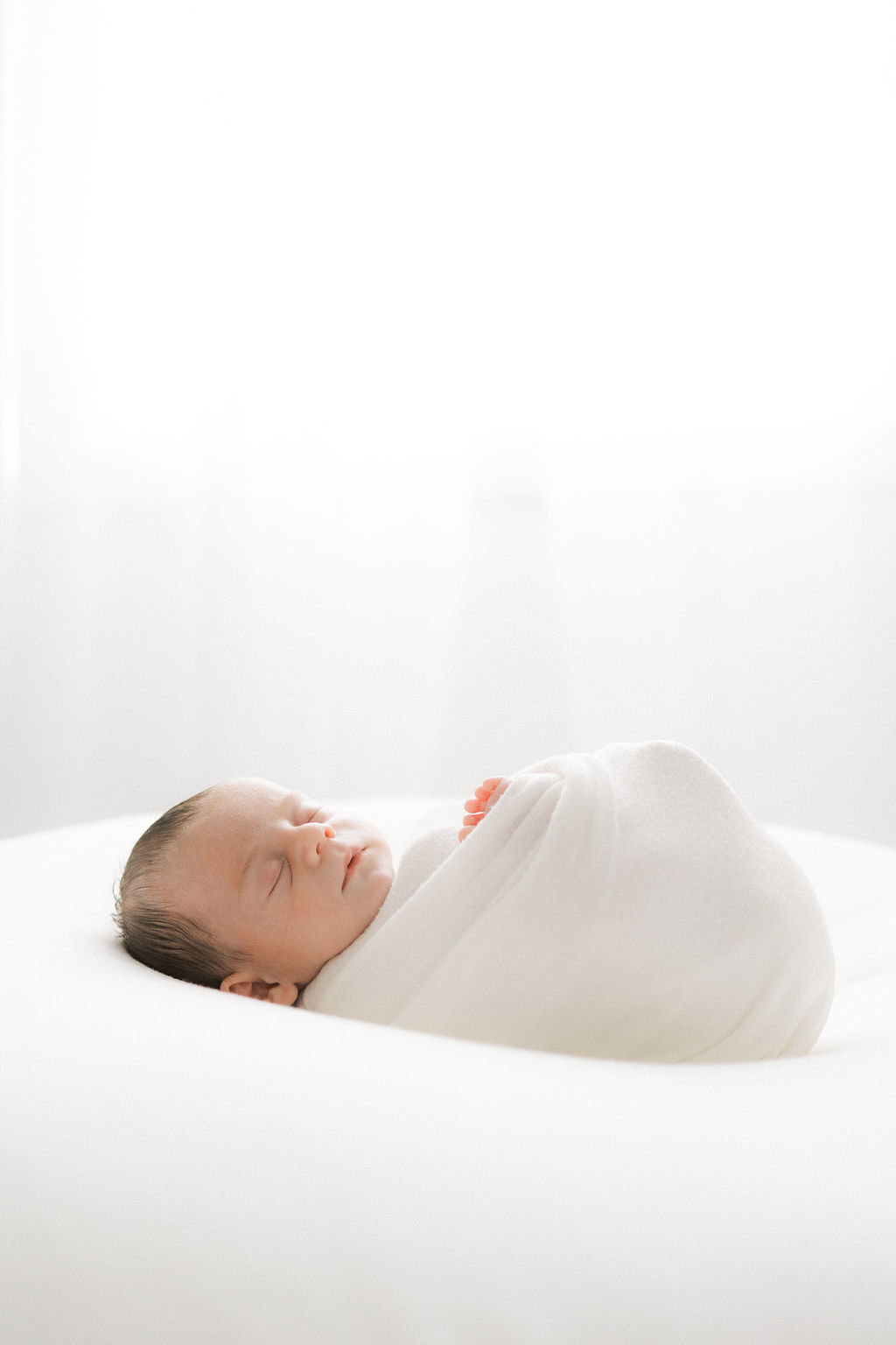 A newborn baby sleeps in a swaddle on a white bed in front of a window