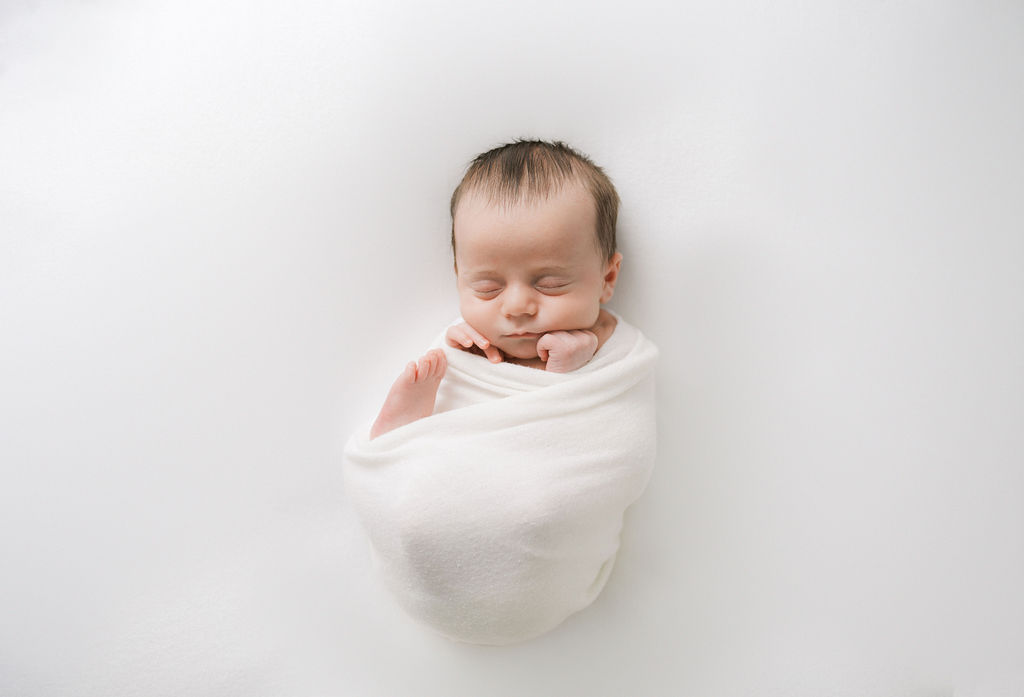 A newborn baby sleeps in a white swaddle with one foot sticking out the top purple bow teaneck