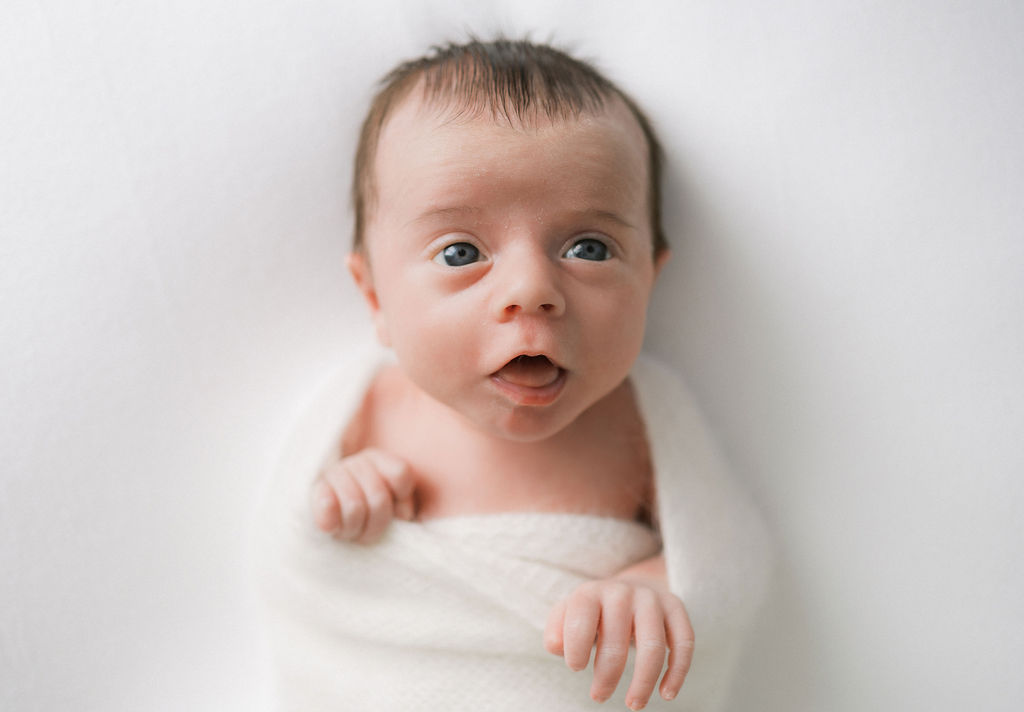 A newborn baby lays in a white swaddle with mouth and eyes open purple bow teaneck