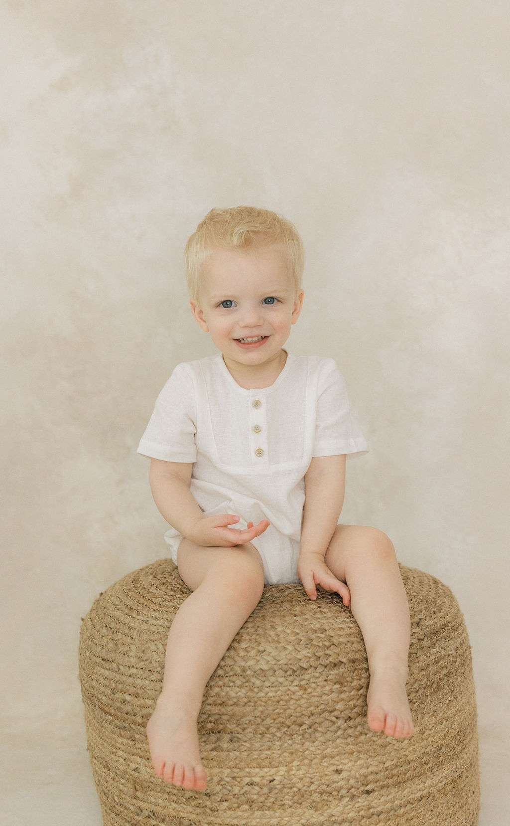 A young toddler in a white onesie sits on a woven stool iin a studio baby furniture stores nj