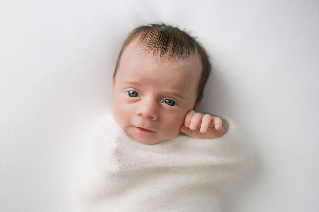 A newborn baby lays in a knit swaddle with one hand sticking out and eyes opeen