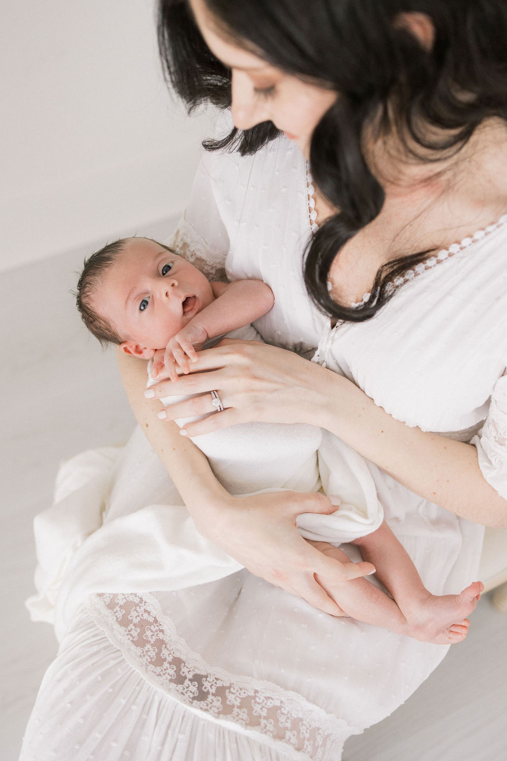 A new mother in a white dress holds her newborn baby in her lap while sitting in a studio cuties boutique