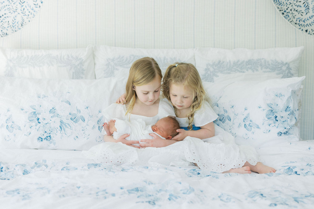 Two young sisters sit on a white and blue floral bed holding their sleeping newborn sibling