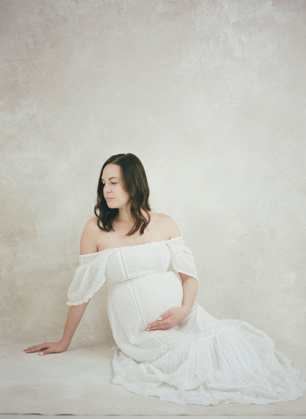 A woman lays across the floor in a studio wearing a white off-the-shoulder maternity dress