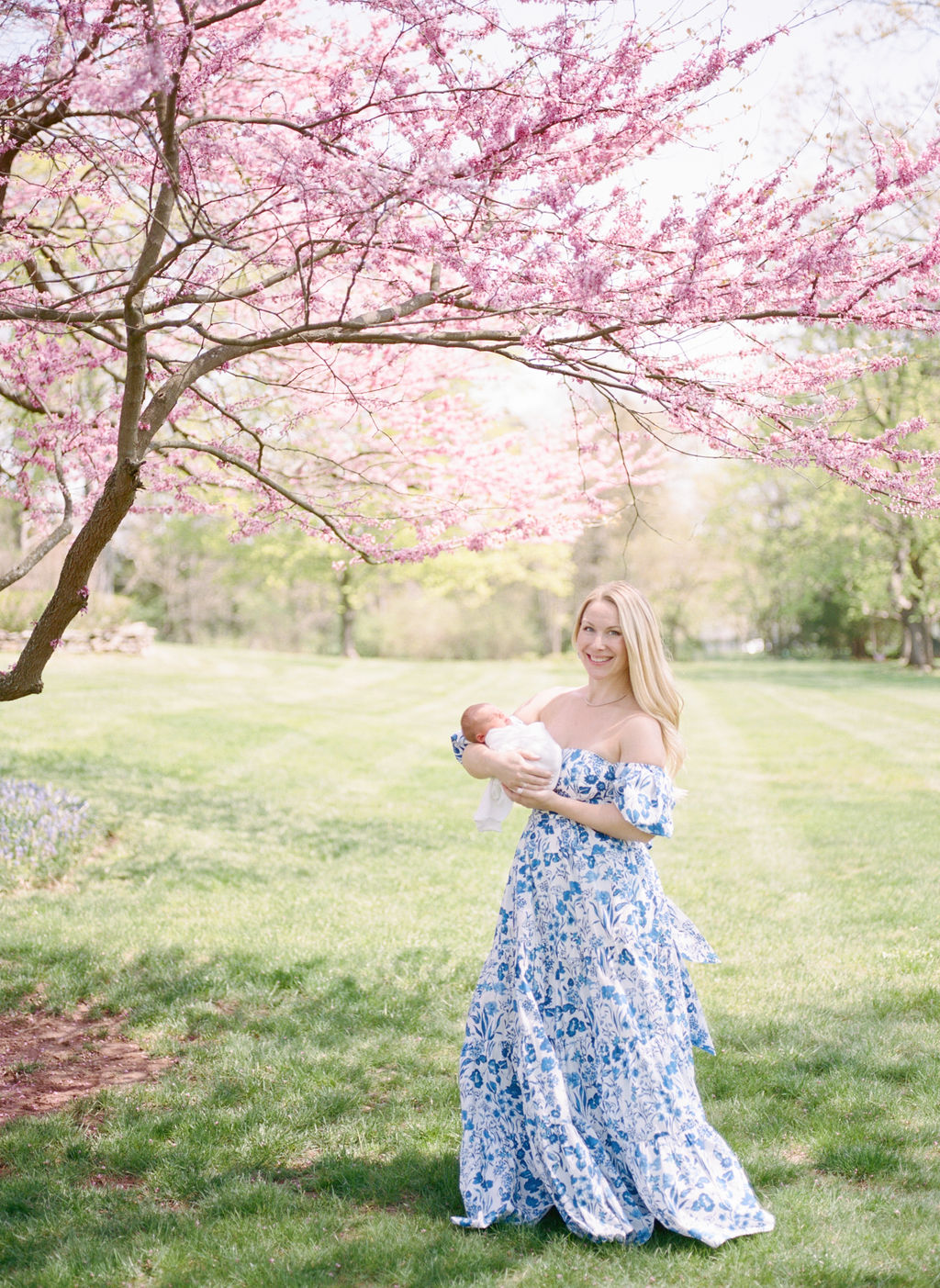 A woman in a blue floral dress stands under a pink blooming tree in a park holding her newborn baby prenatal yoga northern nj