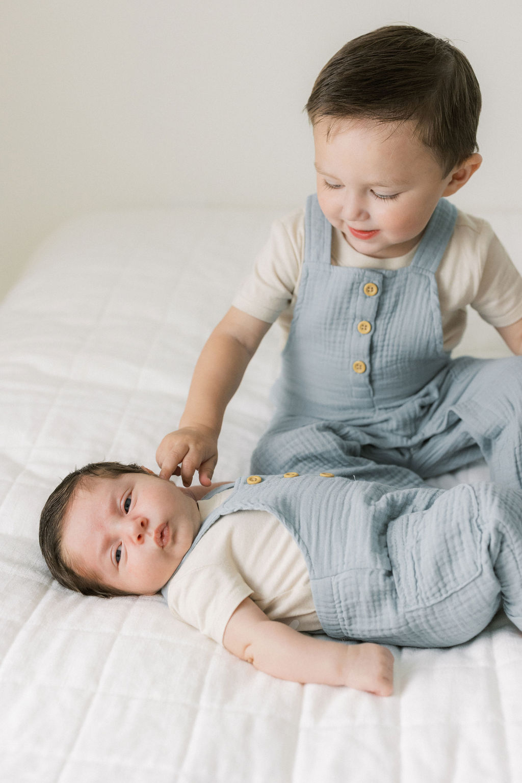 A young toddler boy in blue suspenders touches the cheek of his newborn baby brother laying in a matching suit in front of him