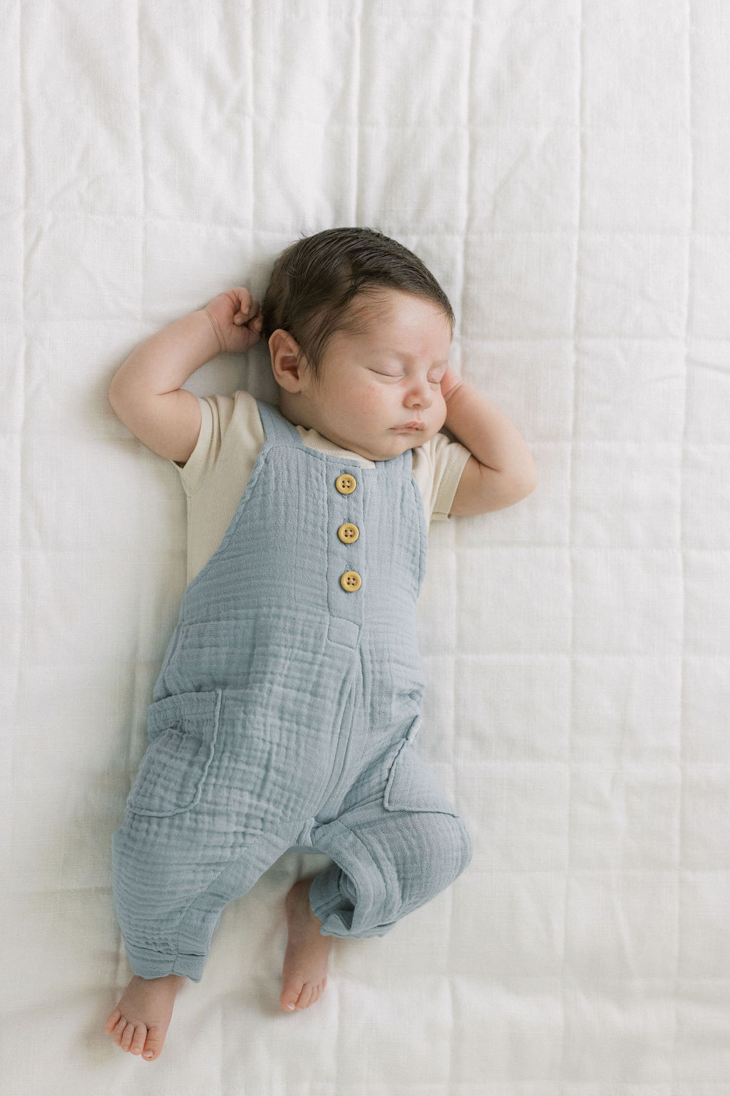 A newborn baby sleeps in blue suspenders on a white bed mommy and me classes nj