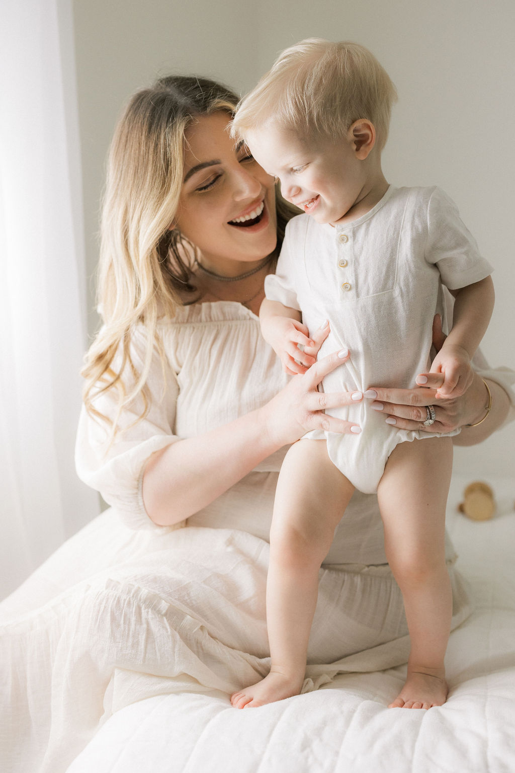 A mother plays with her toddler son in a white onesie and white dress