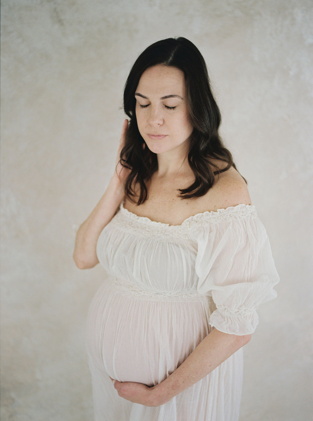 A mom to be in a white maternity dress runs a hand through her hair while standing in a studio
