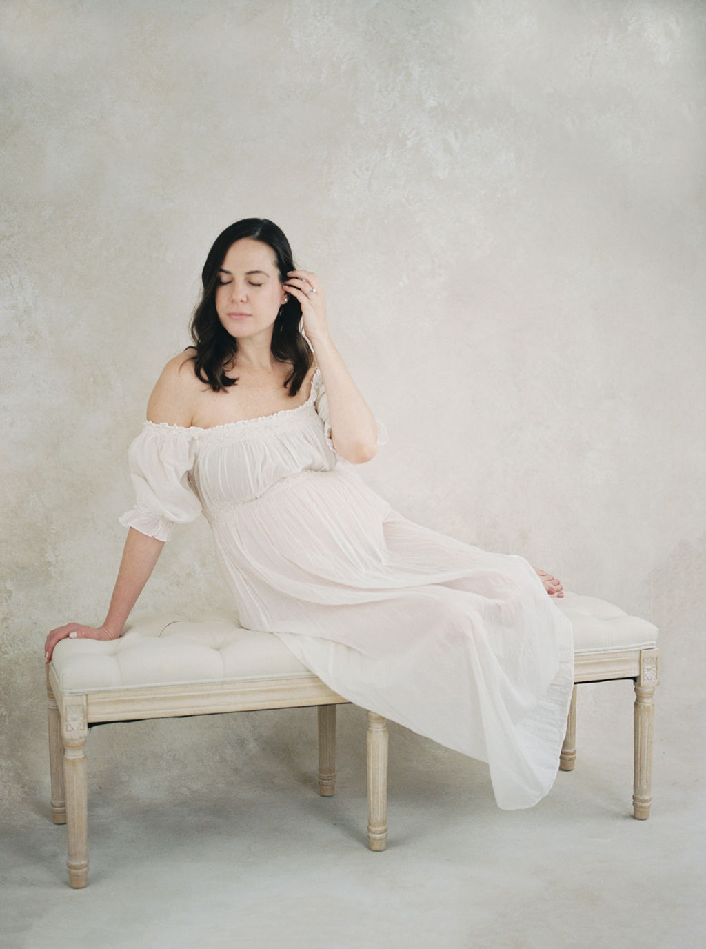 A mother in a white dress sits across a bench in a studio the nesting place nj
